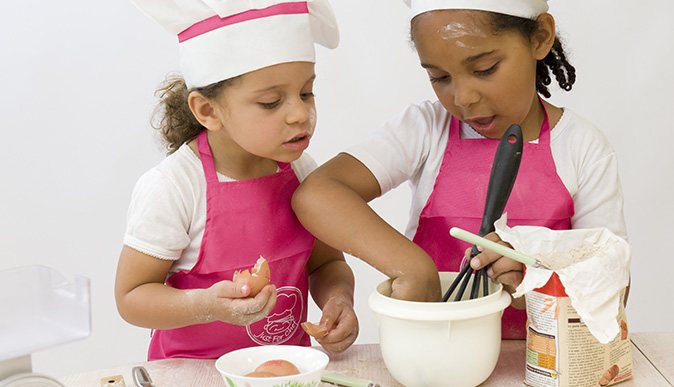 Duas meninas preparando alimentos, uma segurando um ovo e outra com uma colher mexendo numa tigela